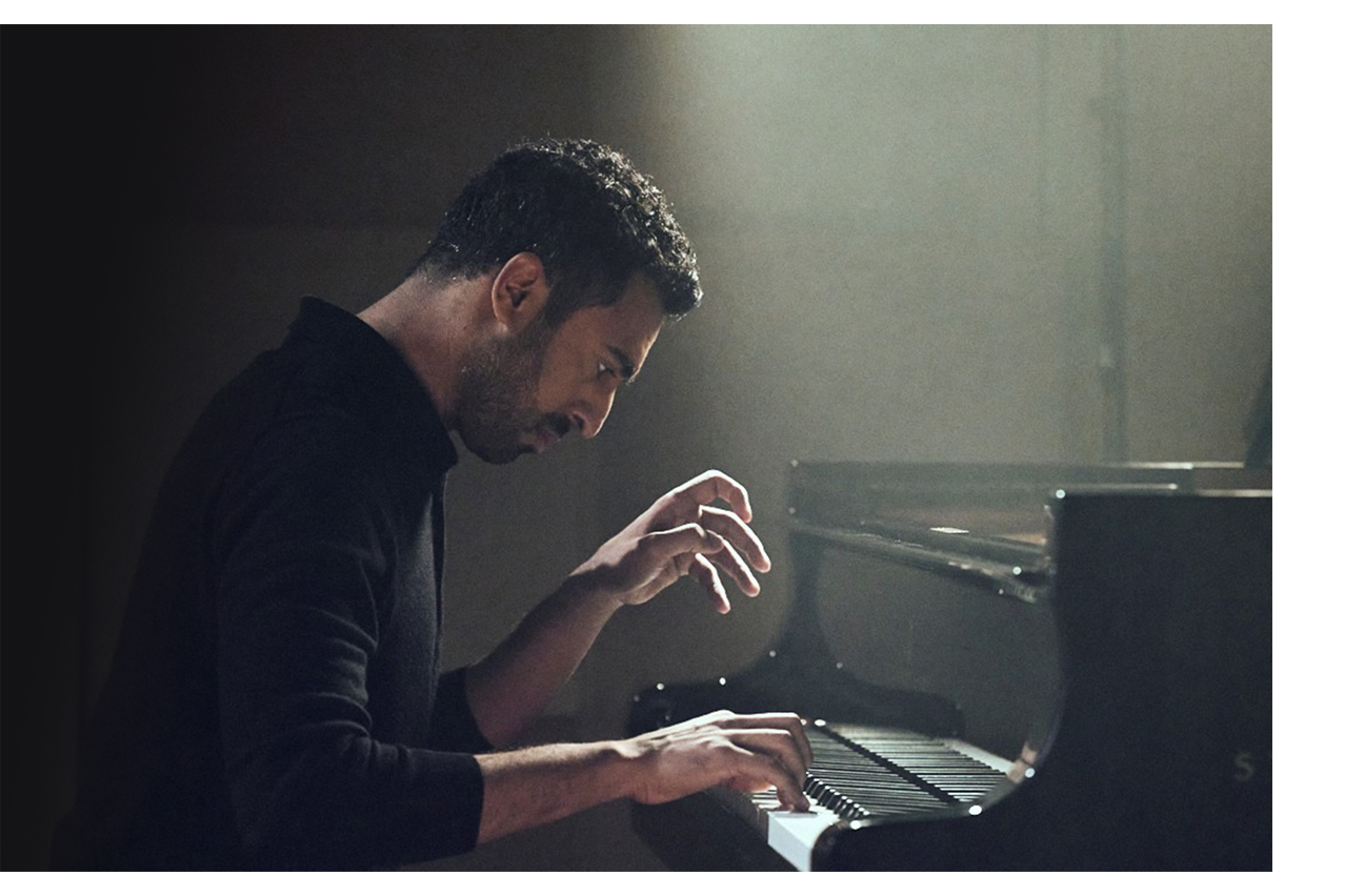 A man wearing a black jumper playing the piano under a spotlight.